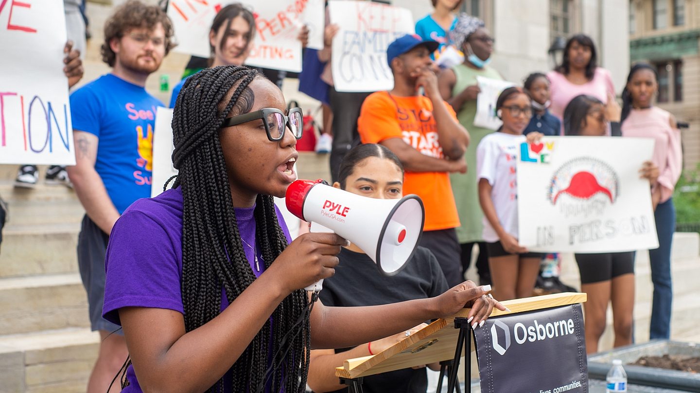 Youth Action Council member speaking through megaphone with a group of rally supporters standing in the background.