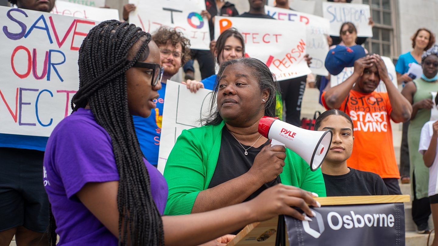 Deputy Brooklyn Borough President Rev. Kim Council holds the megaphone and stand at the podium next to Osborne Youth Action Council member.
