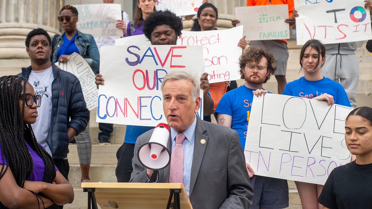 Assemblymember and bill sponsor David I. Weprin stands at the podium in the center of the photo with a megaphone surrounded by YAC members.