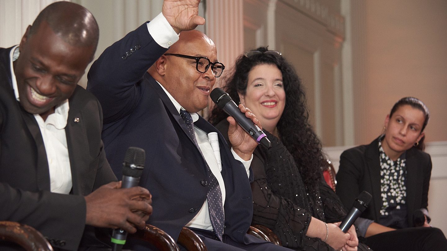 Close-up of panel speakers sitting and smiling at one another, Eddie Gibbs has one hand raised and the other holding a microphone.