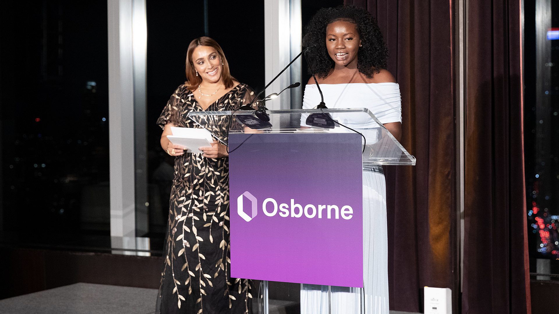 Left to right: Aylyn Restituyo stands to the right of Jamila Small looking at her speak at the podium.