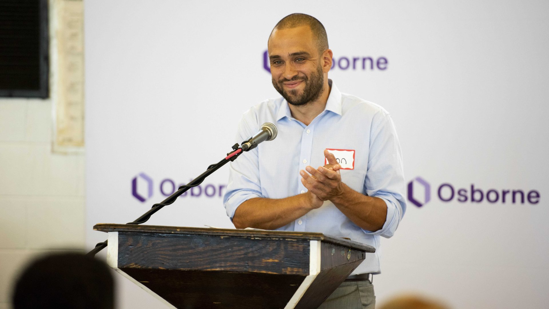 Jonathan Monsalve smiles while standing at the podium at a graduation inside a prison. His hands are clasped together in a clap.