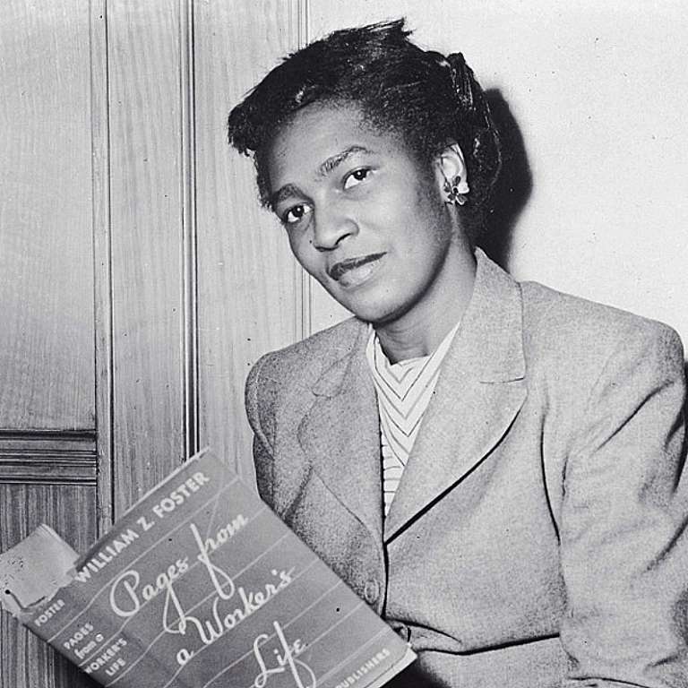 Photo of Claudia Jones sitting and reading a book with her face turned towards the camera.